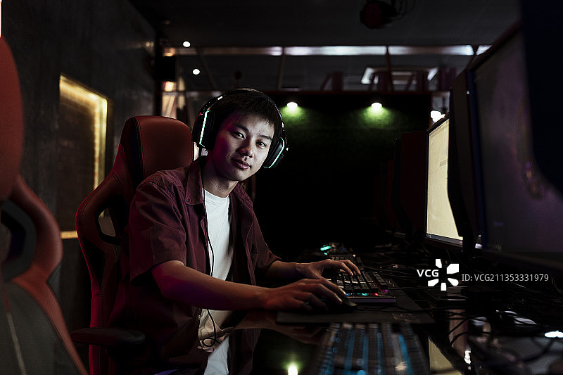 年轻的亚洲青少年玩家戴着耳机坐在红椅子在网吧。图片素材