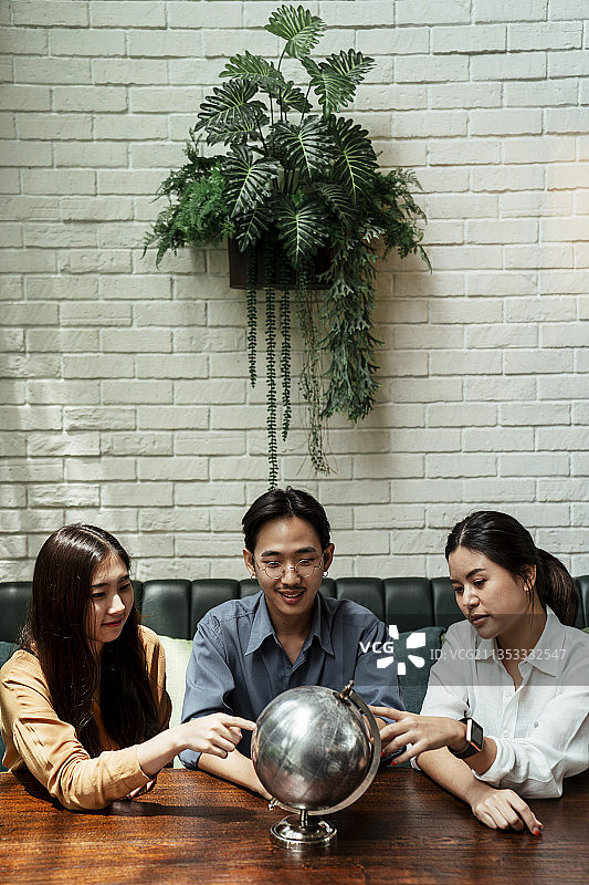 三个亚洲年轻人指着金属地球仪，坐在一起，选择他们想去的国家。图片素材