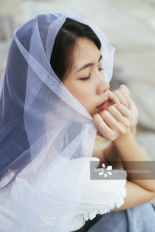 近距离-年轻的亚洲长发女子新娘的肖像与白色面纱坐在户外的海滩上。图片素材