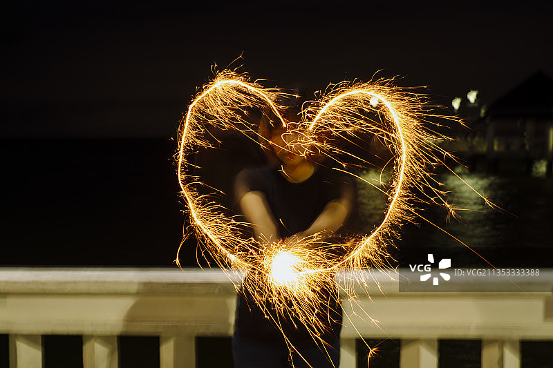 一个女人在晚上用烟火画出一个心形。长时间的曝光。在海滩上。图片素材