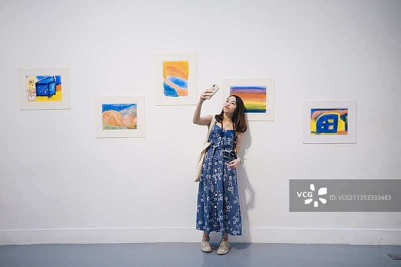 漂亮的时髦亚洲女人在画廊里穿着蓝色的衣服，用智能手机自拍。图片素材