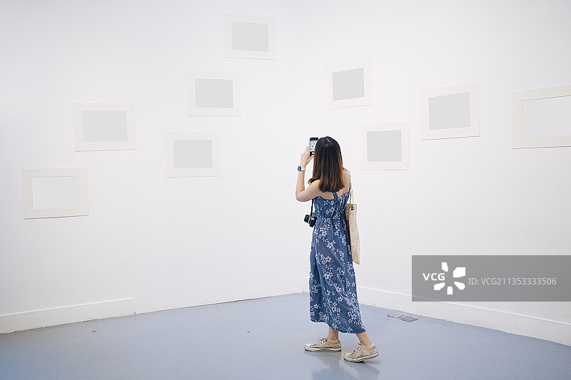年轻的亚洲泰国妇女在蓝色的衣服拍照与智能手机在白色画廊。图片素材