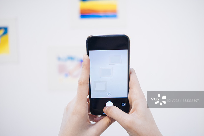 在白色画廊中，手持智能手机的女性正在拍摄艺术品。图片素材