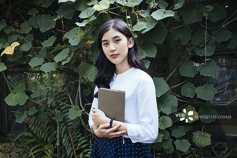 美丽的亚洲女人在热带花园拿着一本书与她的手，站在活墙外的石头房子。女孩和书。小说作家。图片素材