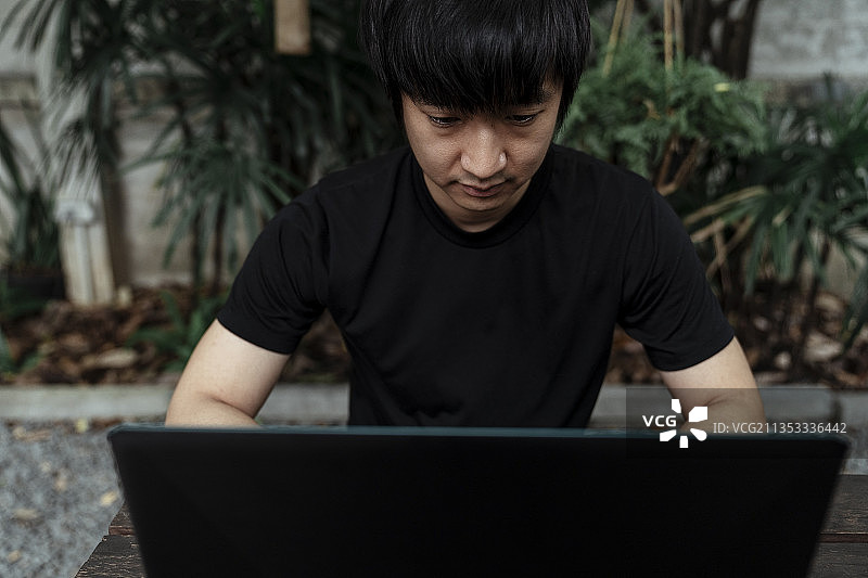 一个穿着黑色t恤的男人正坐在花园里分析他笔记本电脑里的信息。图片素材