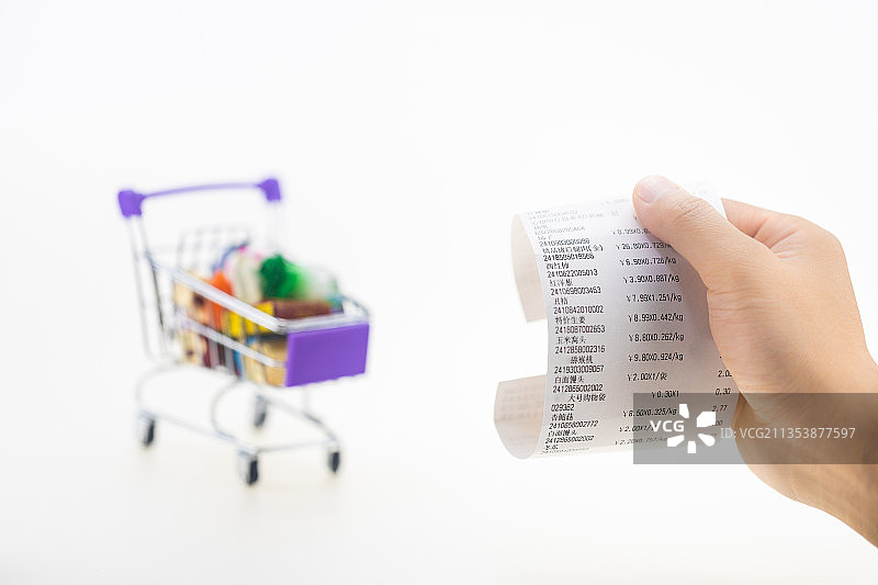 白色背景里的购物车前，男性的手里拿着一张超市小票图片素材