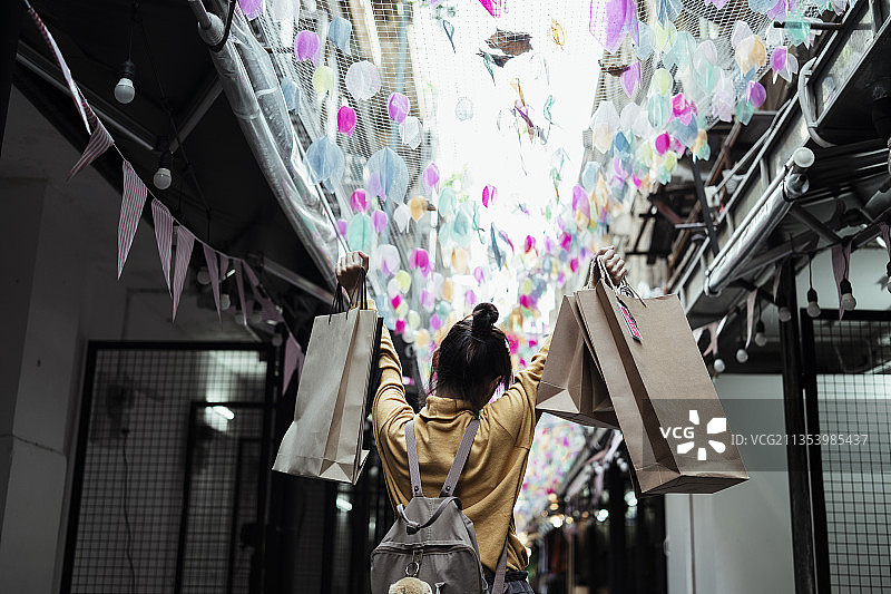 后视图-亚洲妇女穿着黄色毛衣拿着购物纸袋在狂欢节活动。购物爱好者的女孩。图片素材