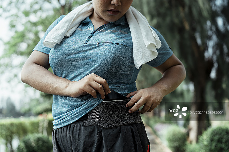 胖胖的运动亚洲女人穿着蓝色运动服从小口袋里拿智能手机在公园。图片素材