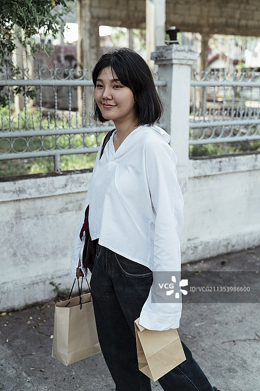 年轻的亚洲女人穿着白衬衫，拿着购物纸袋走在街上。图片素材