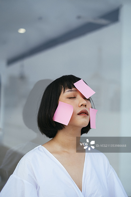 一个黑色短发的女人坐在她的玻璃办公室里，身上贴着很多便利贴。图片素材