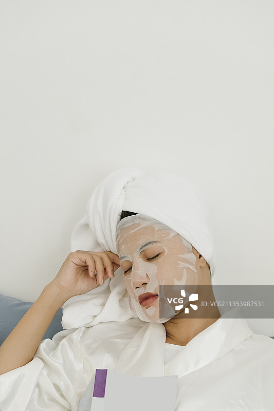 年轻女子穿着白色浴衣敷着化妆面具躺在客厅的沙发上。图片素材