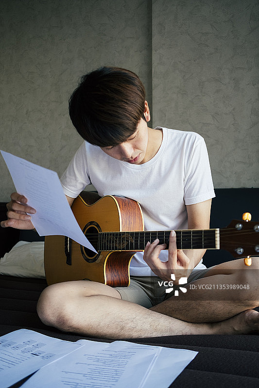 年轻的泰国吉他手练习原声吉他和阅读抒情纸笔记在卧室。图片素材