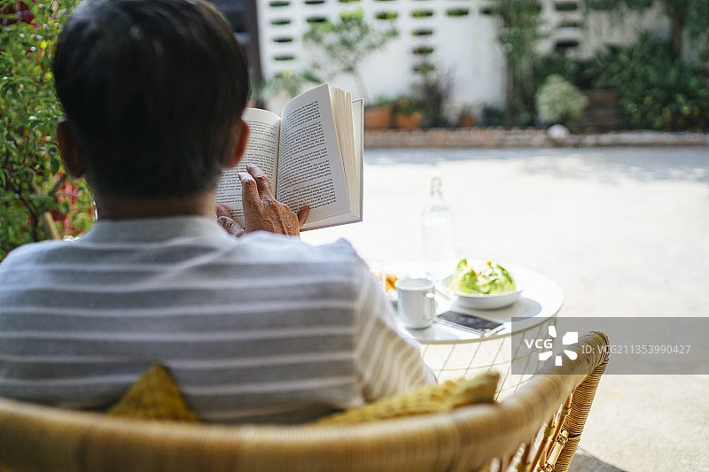 一个老人在他的院子里吃完早餐后正在看书，越过肩膀射击。图片素材