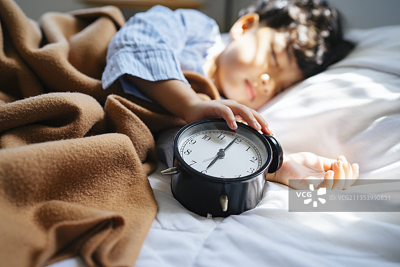 白天躺在床上时，男孩手里拿着闹钟的裁剪图像。图片素材