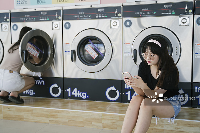 戴着眼镜的长发女孩在等待洗衣机的时候使用智能手机。图片素材