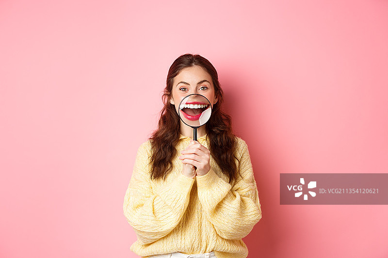 在粉色背景下，透过放大镜微笑的快乐女人图片素材
