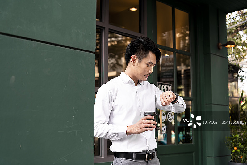 成年亚洲泰国商人在白衬衫上拿着一杯咖啡，看着手上的手表检查时间。图片素材