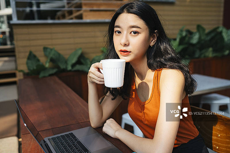 亚洲美女穿着橙色衬衫与笔记本电脑工作在咖啡馆在城市的咖啡馆。图片素材