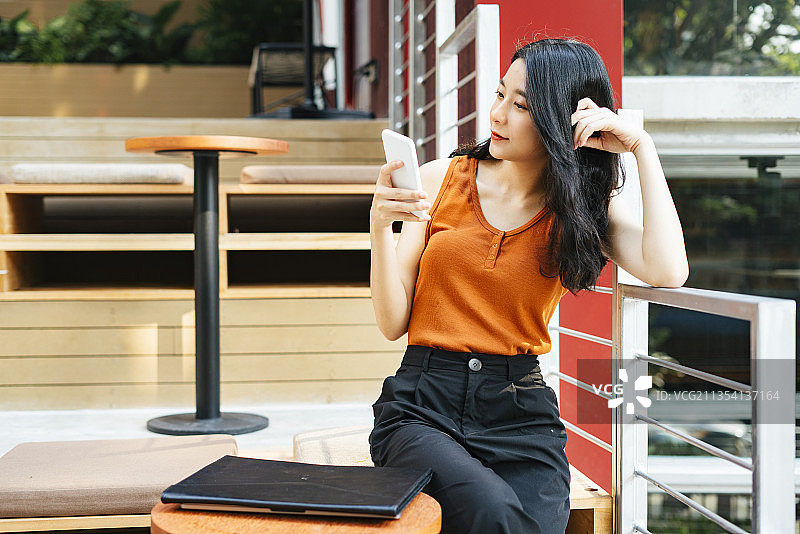 性感的亚洲美女穿着橙色衬衫，拿着智能手机坐在户外工作空间。图片素材
