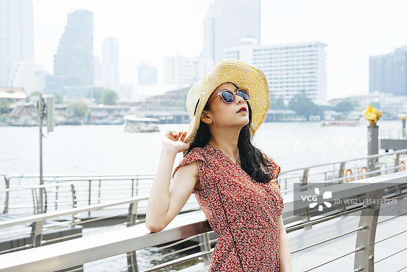 年轻美丽的泰国亚洲旅行者女人穿着红色的裙子，帽子，和太阳眼镜在曼谷的一个码头上旅行。图片素材