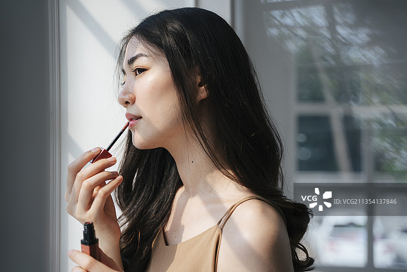 波浪形长发女孩站在窗边用唇刷涂口红。图片素材