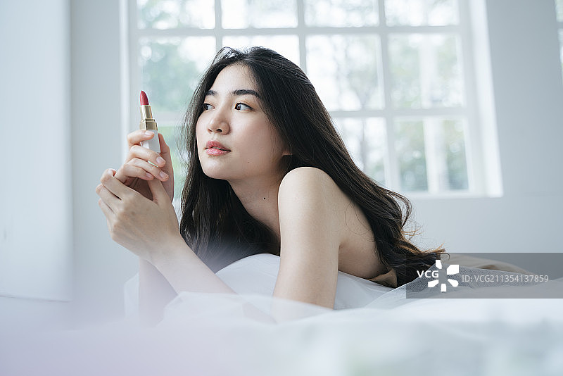 卷长发的女人早上趴在床上，拿着口红，然后转动它。图片素材