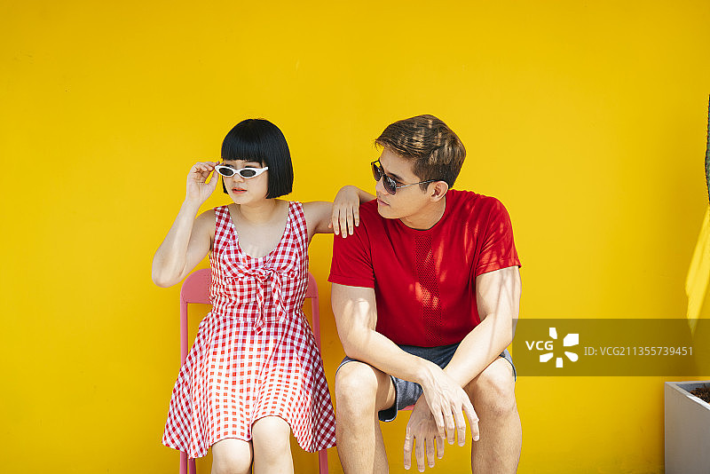 一对亚洲的泰国夫妇穿着红色衬衫和太阳镜坐在长凳上的暑假在橘黄色的背景。图片素材