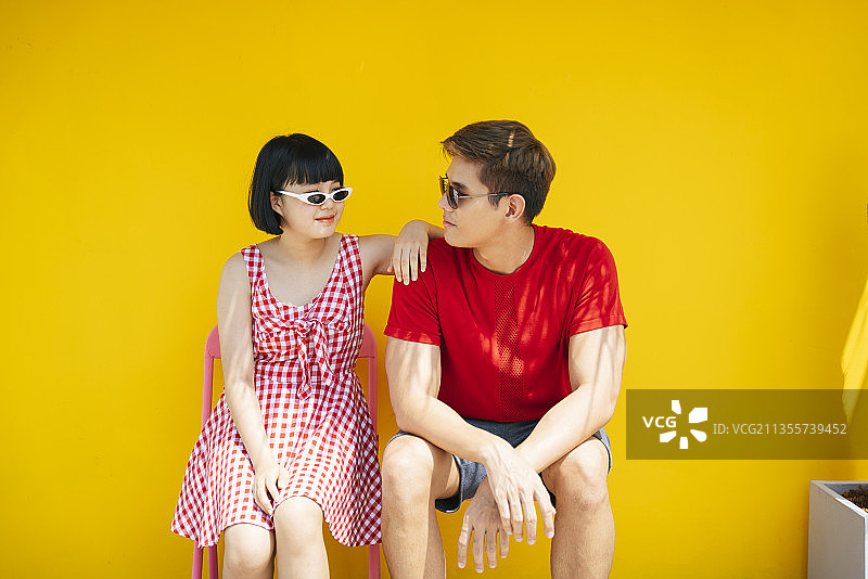 一对亚洲的泰国夫妇穿着红色衬衫和太阳镜坐在长凳上的暑假在橘黄色的背景。图片素材