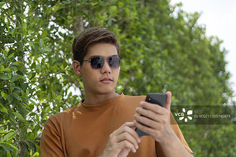 年轻英俊的亚洲泰国男子戴着太阳镜使用智能手机在自然。图片素材