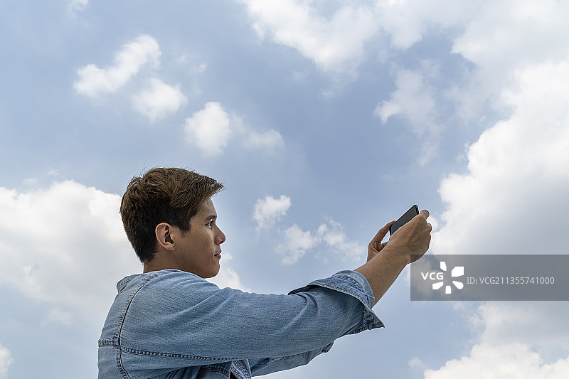 亚洲泰国当地男子穿着牛仔夹克在蓝天下使用智能手机。图片素材