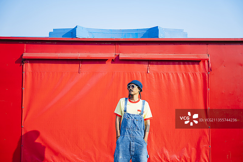一个穿着黄色t恤和牛仔工装裤的男人站在红色的拖车前踢着车。图片素材