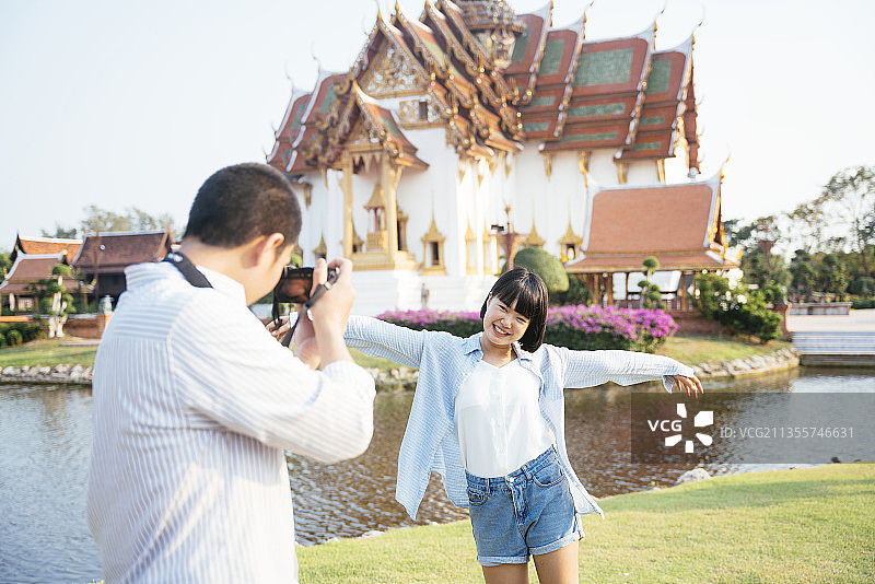 一个亚洲男朋友在河边的佛寺用相机给他的女朋友拍照。图片素材