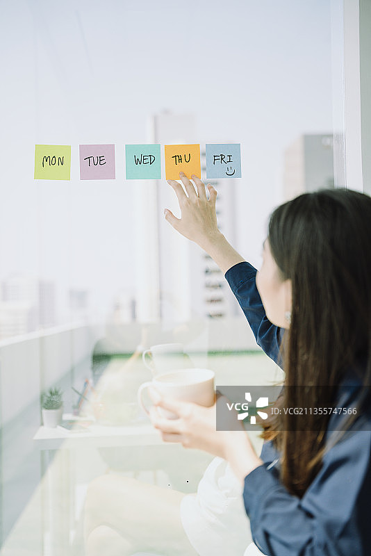 美丽的亚洲年轻的泰国女人长黑头发制作时间表在粘纸笔记在玻璃墙上。商人安排会议。图片素材