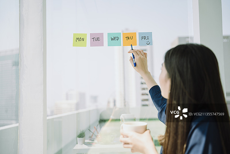 美丽的亚洲年轻的泰国女人长黑头发制作时间表在粘纸笔记在玻璃墙上。商人安排会议。图片素材