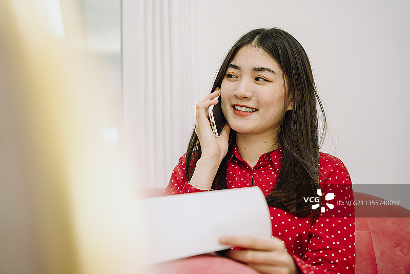 漂亮的亚洲泰国长黑发女人在红色衬衫坐在粉红色的沙发沙发上使用智能手机与客户联系和检查清单上的文件工作。居家办公在家工作。图片素材