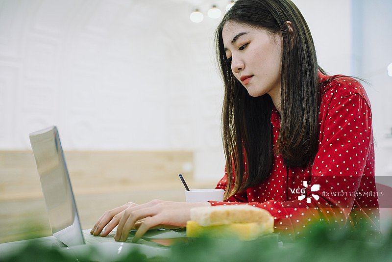 漂亮的亚洲泰国长黑头发女人在红色衬衫工作在笔记本电脑在家里。在家工作的概念。图片素材