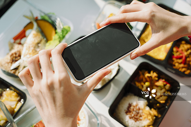 前视图。一个女人用智能手机拍照。饭盒放在桌上。图片素材