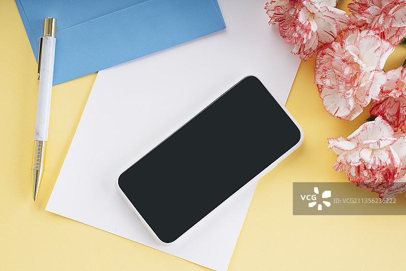 模拟婚礼模板。智能手机、白纸、蓝色信封和黄色背景上的康乃馨。副本的空间。图片素材