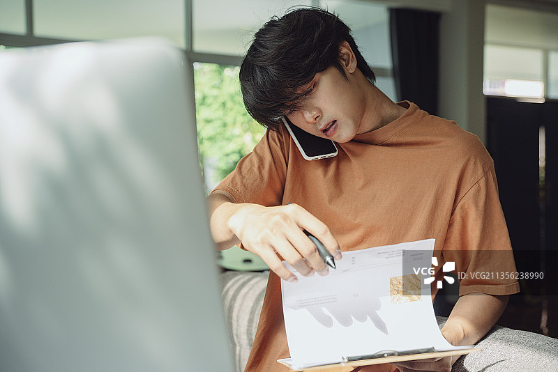 忙碌的亚洲泰国商人在家工作，一边和同事打电话一边看文件。业务分析。图片素材
