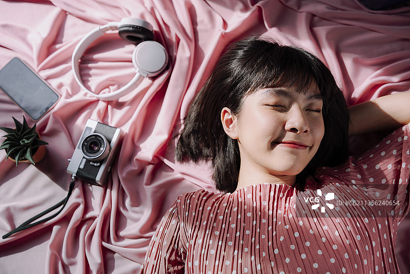 俯视图-美丽的亚洲泰国女人短黑发睡在粉红色的背景。摄像头,耳机,智能手机。旅行的梦想家。图片素材