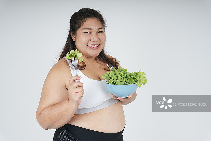 开朗快乐的亚洲胖女人在白色运动胸罩享受吃沙拉碗在白色的背景。图片素材