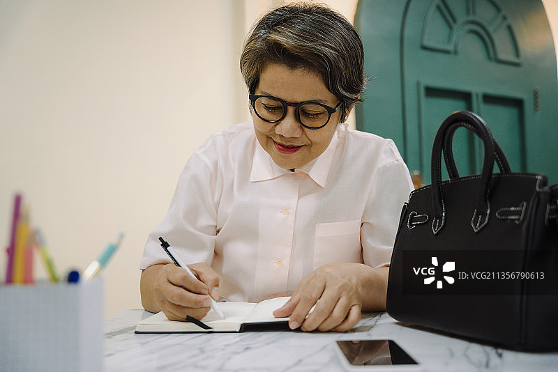 亚洲老妇人戴着眼镜在书上写日记。图片素材