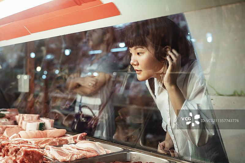 泰国黑头短发女子身穿白衬衫，看着猪肉放在弯曲的玻璃陈列柜里，啪地一声穿过玻璃。图片素材