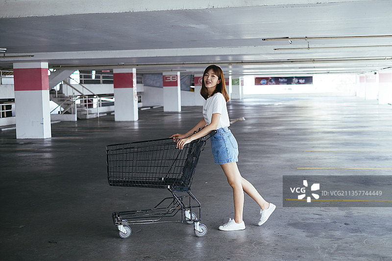 年轻的泰国亚裔女子穿着白衬衫推着购物车在停车场。图片素材