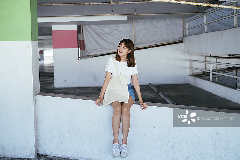 年轻的泰国亚裔妇女坐在停车场，肩上背着购物布袋。图片素材