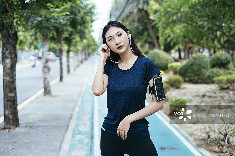 运动的亚洲妇女在公园的跑道上，穿着深色运动服，戴着耳机听着音乐。图片素材