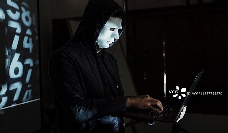 黑客穿着连帽衫，戴着白色面具入侵系统，笔记本电脑在暗室服务器。图片素材