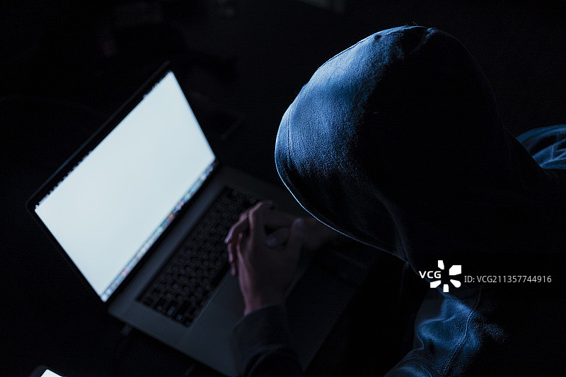 笔记本电脑屏幕上的拷贝空间-黑色连帽衫黑客在暗室用笔记本电脑入侵电脑网络。网络安全的概念。图片素材