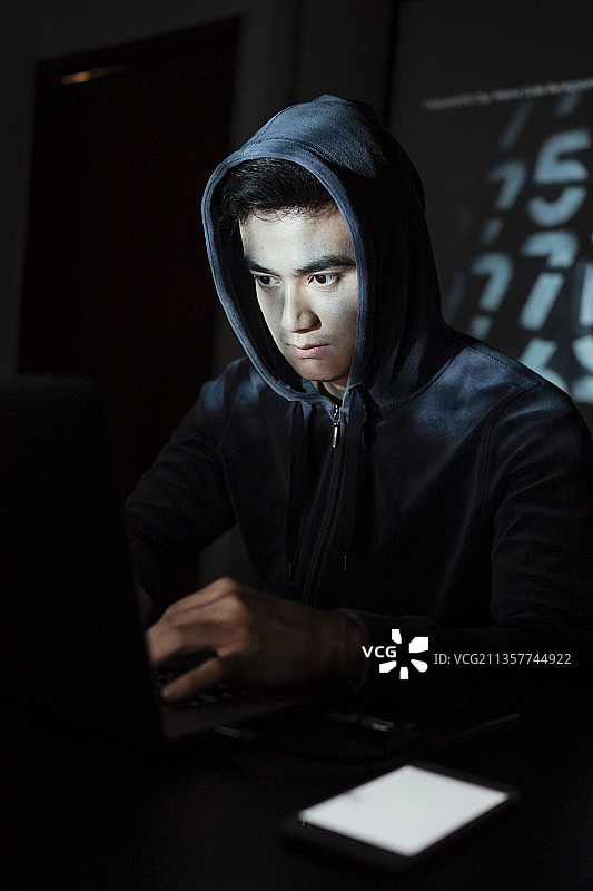 亚洲黑客戴着黑帽在暗室里用笔记本电脑和智能手机入侵政府系统。图片素材