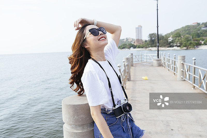 年轻的亚洲旅行者泰国女摄影师穿着白色的裙子和太阳镜站在海上码头。图片素材